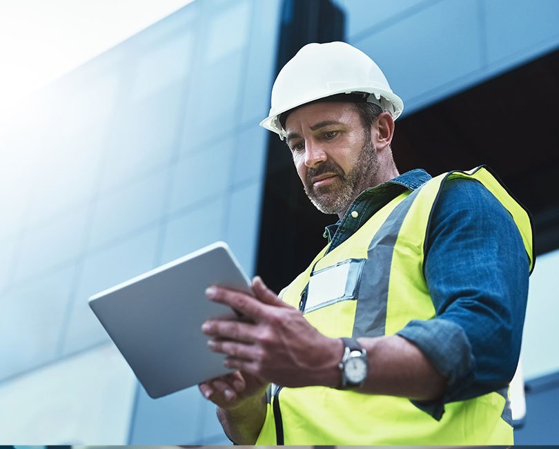 A person wearing a white hard hat and a high-visibility safety vest is looking at a tablet. They appear to be an AE firm professional on a construction site with a modern building in the background. The sky is clear and bright.