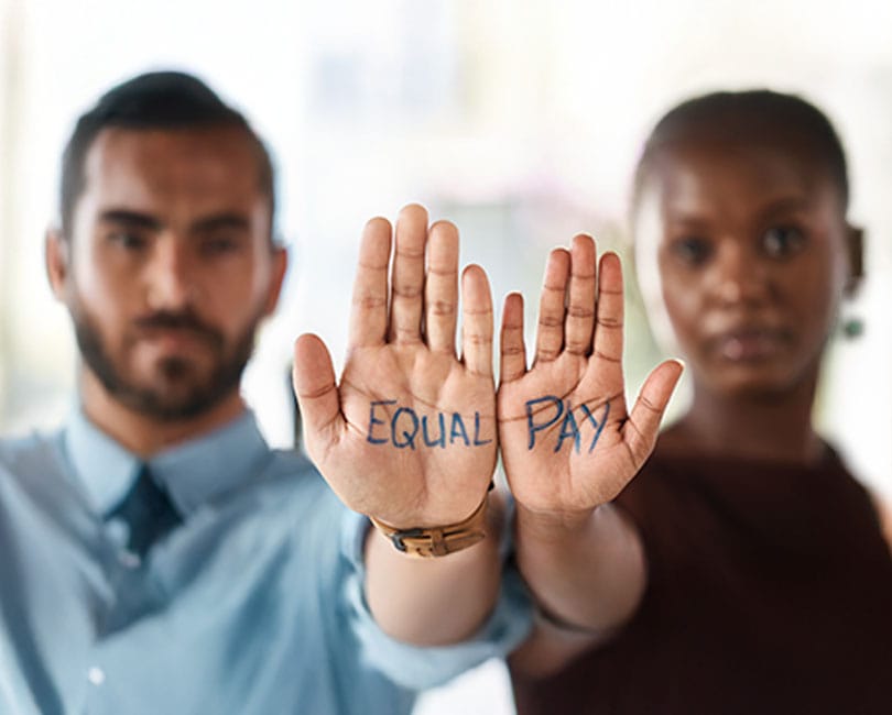 Two people hold up their hands with "Equal Pay" written on their palms, blurred faces in the background.