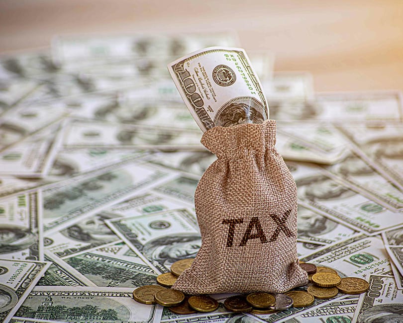 A burlap sack labeled "TAX" stands upright, stuffed with a rolled-up U.S. hundred-dollar bill, surrounded by scattered coins and piles of U.S. dollar bills. The scene represents financial and tax-related concepts.