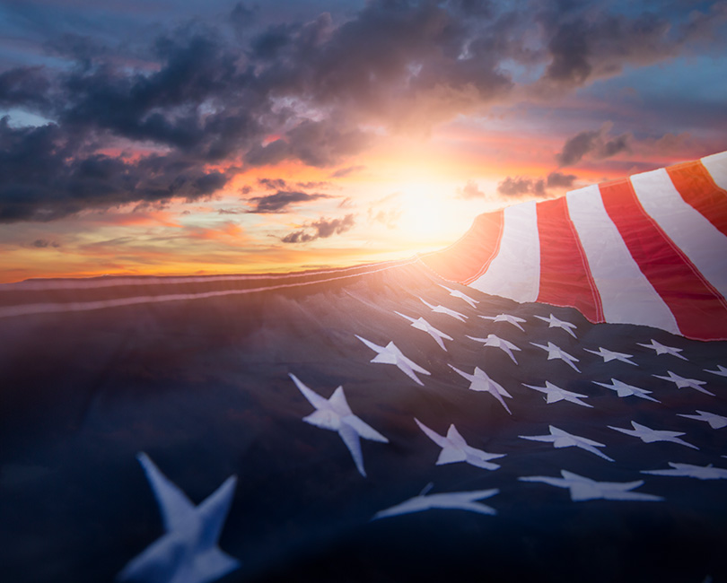 An American flag waves against a dramatic sunset sky with dark clouds and a burst of sunlight.