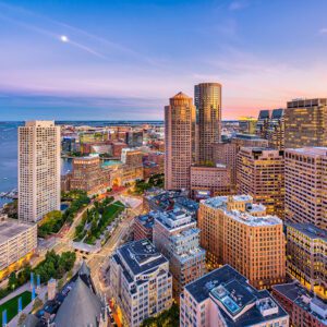 Aerial view of Boston, Massachusetts at sunset, showcasing a cityscape where modern skyscrapers meet historic buildings. The waterfront adds charm as the sky melds blue and orange hues, dotted with a few scattered clouds.