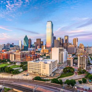 Aerial view of the Dallas, Texas skyline with diverse buildings under a clear blue sky. In the foreground, multiple train tracks intersect. The tallest building reflects the pastel colors of sunset, enhancing the urban landscape.