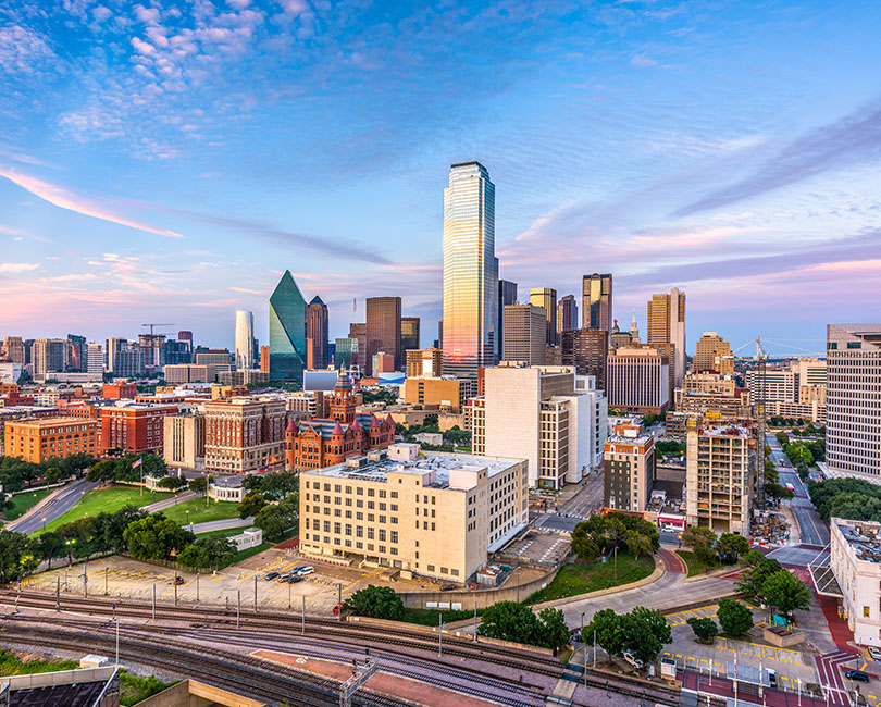 Aerial view of the Dallas, Texas skyline with diverse buildings under a clear blue sky. In the foreground, multiple train tracks intersect. The tallest building reflects the pastel colors of sunset, enhancing the urban landscape.