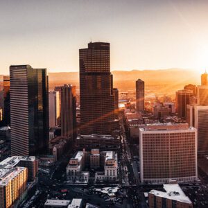 Aerial view of the Denver, Colorado skyline at sunset with tall skyscrapers casting long shadows. The sun is low on the horizon, creating a warm glow. Snow dusts the rooftops of several shorter buildings, and mountains rise majestically in the distance.