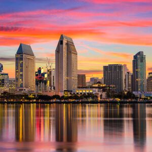 San Diego's vibrant city skyline reflects on calm water during sunset. Tall buildings, including a distinctive triangular-roofed structure, stand against a colorful sky with shades of orange, pink, and purple.