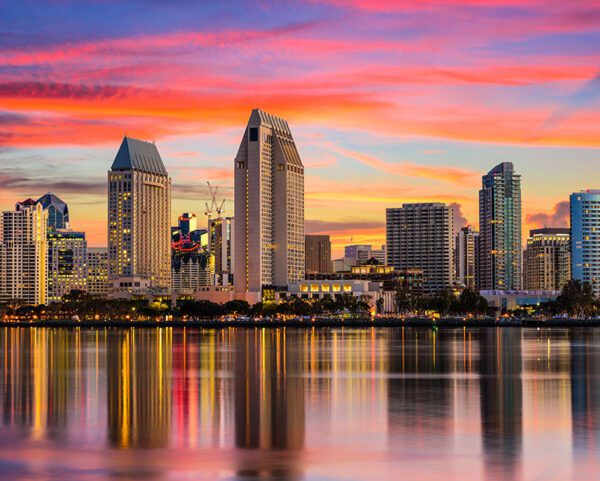 San Diego's vibrant city skyline reflects on calm water during sunset. Tall buildings, including a distinctive triangular-roofed structure, stand against a colorful sky with shades of orange, pink, and purple.