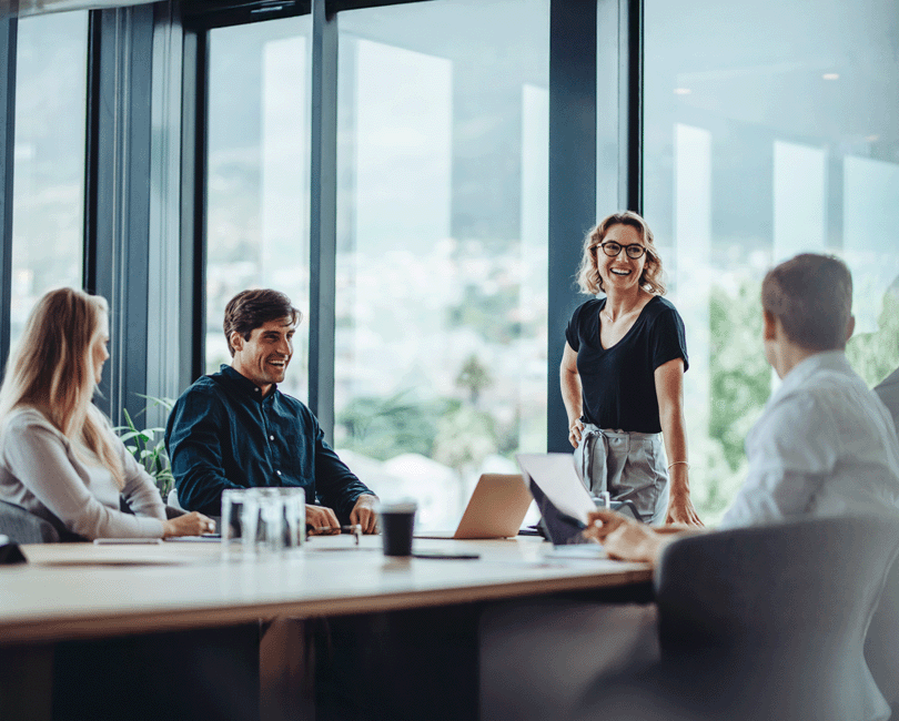 A woman stands, smiling, at the head of a conference table during an M&A meeting. Three colleagues are seated, engaged in conversation. The room has large windows with a view of a cityscape.